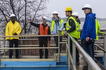 A group in hard hats and yellow vests points at something in the distance while standing at a wastewater treatment plant.