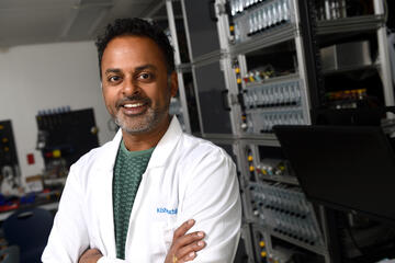 A man in a white lab coat poses with his arms crossed in front of him and smiles